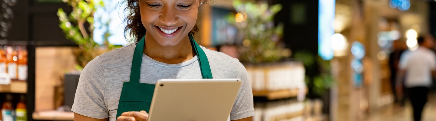en person som står med en tablet, med en uskarpet bakgrunn som viser blomster og og noen mat produkter